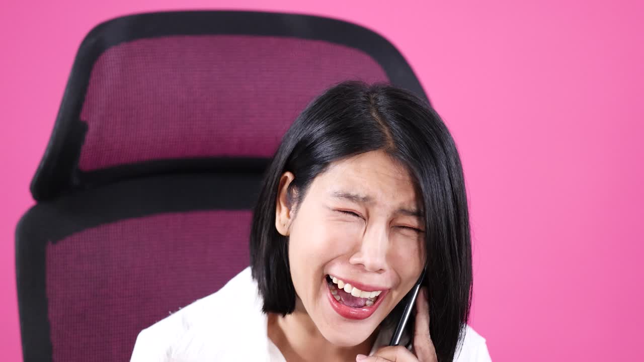 Asian woman in office chair expresses surprise, excitement, and joy on phone against pink background