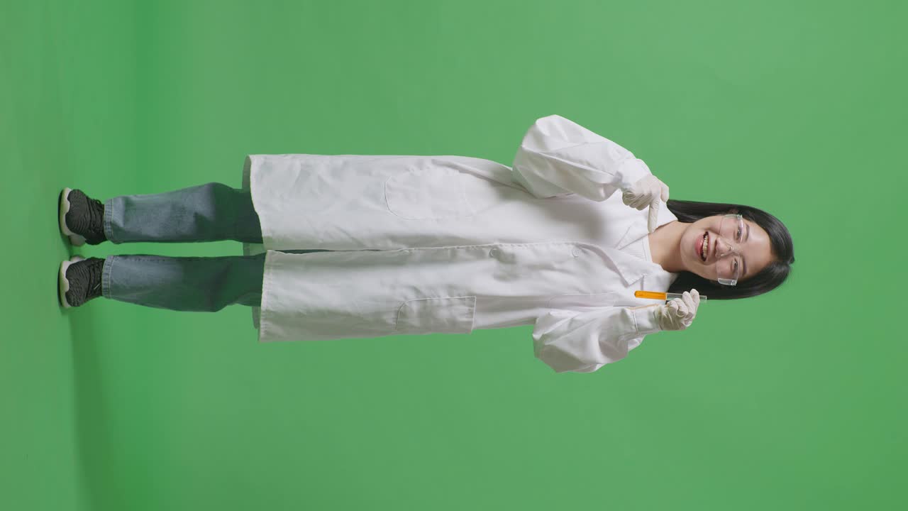 Full Body Of Asian Woman Scientist Smiling And Pointing To Orange Liquid In The Test Tube In Her Hand While Standing On The Green Screen Background In The Laboratory