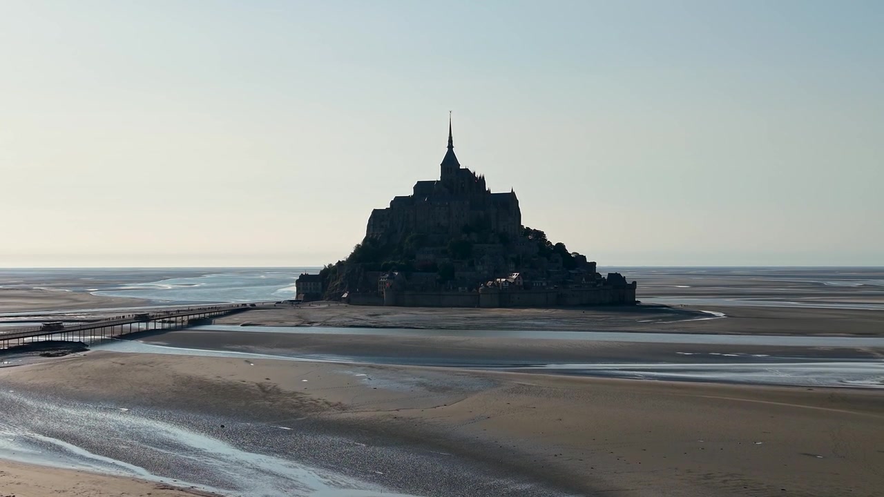 Bredt filmisk luftfoto fra drone af Mont Saint-Michel omgivet af store vadehavsflader ved lavvande. Det ikoniske middelalderkloster rejser sig dramatisk over bugten.