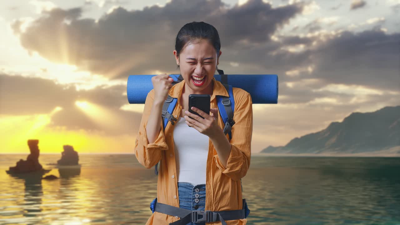 登山用バックパックを背負ったアジア系の女性ハイカーが、スマートフォンを見て目標を叫び、夕暮れ時のビーチに立って成功を祝っています。