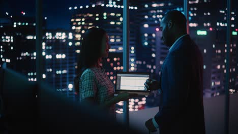 financial analyst talking to investment banker in late evening at work. chatting about business project data on laptop computer. businesspeople have a meeting in broker agency city office.