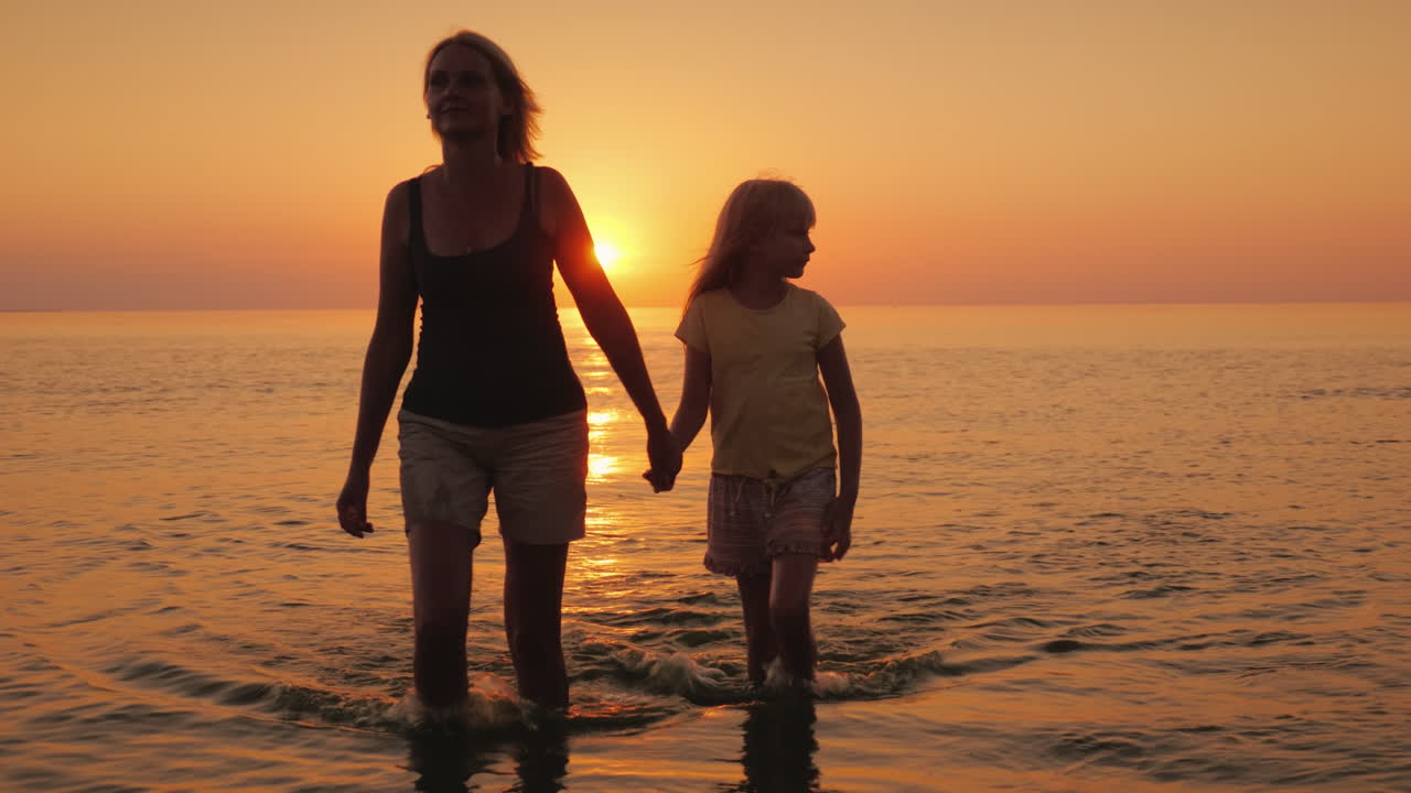 Uma Mãe Atraente Caminha Com A Filha Na Água Tendo O Pôr Do Sol Como Cenário