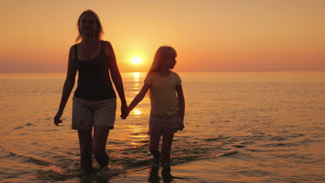 an attractive mother walks with her daughter on the water against the backdrop of the setting sun a