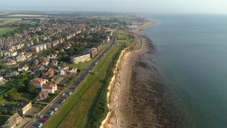 Hoher-Drohnenschuss-Aus-Der-Luft,-Der-Die-Linie-Der-Alten-Hunstanton-Klippen-Mit-Paralleler-Straße-Und-Sandigem-Und-Felsigem-Strand-Hinunterfliegt,-Wird-Das-Meerwasser-Mit-Neuem-Hunstanton-In-Der-Ferne-Im-Norden-Von-Norfolk,-Großbritannien,-Beruhigen