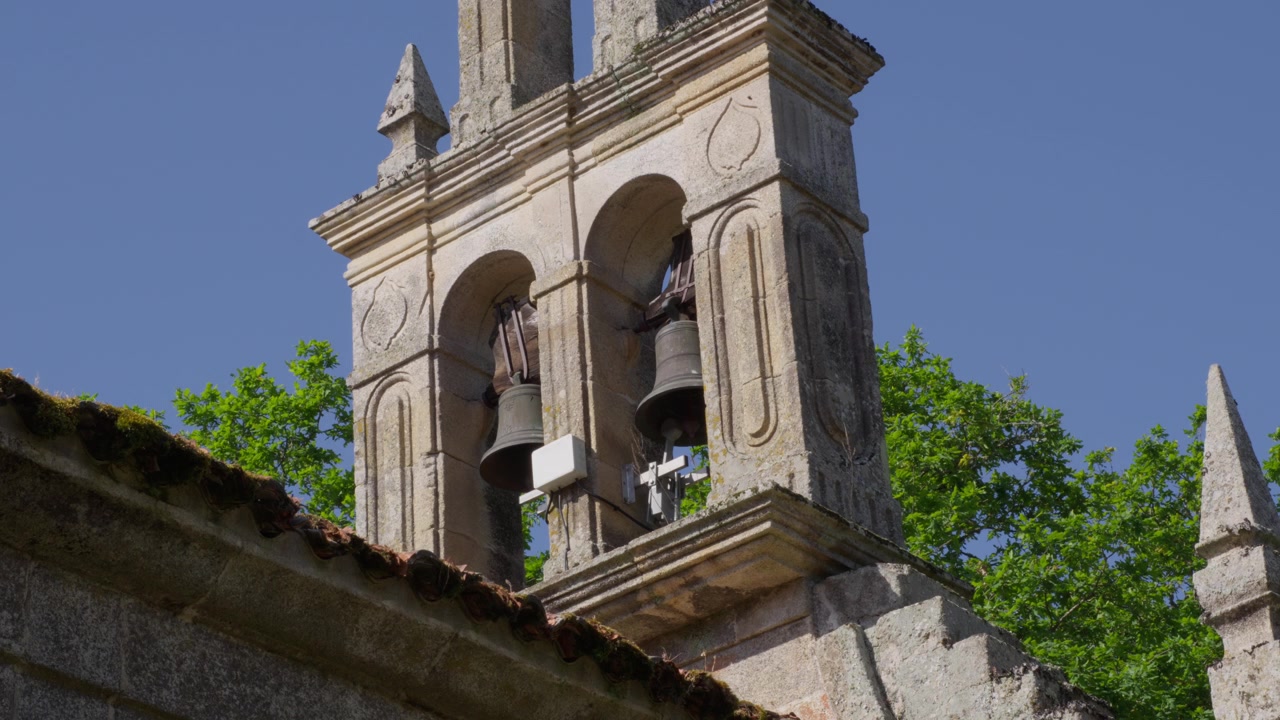 Inquadratura ampia ravvicinata di un vecchio campanile in pietra di una chiesa con dettagli ornamentali, illuminato dal sole contro un cielo blu vibrante e alberi verdi e frondosi. Nessuno, atmosfera serena.
