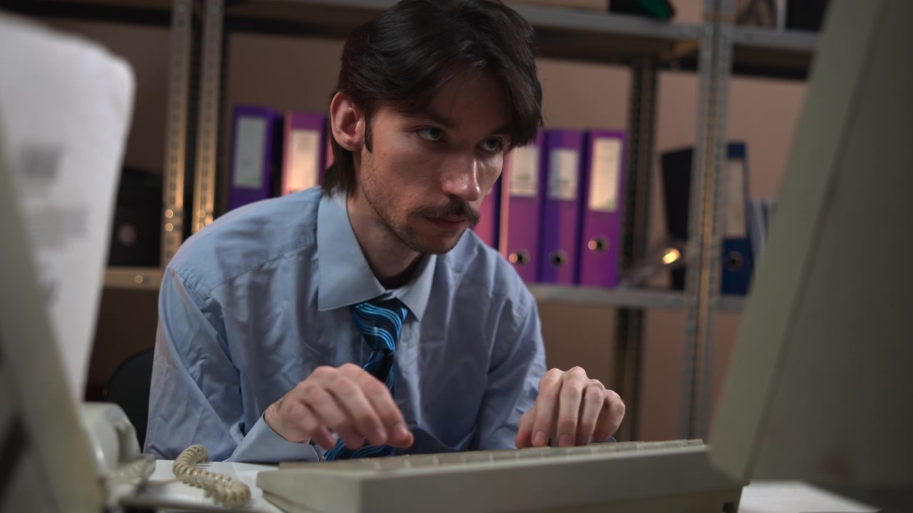 Un uomo serio siede a una scrivania e si china verso una tastiera in un interno d'ufficio poco illuminato, con scaffali di raccoglitori e attrezzature informatiche che creano un ambiente professionale dinamico.