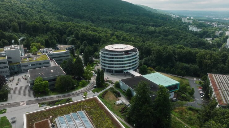 Um drone faz um movimento panorâmico da esquerda para a direita, mantendo o edifício redondo do DKFZ centralizado, revelando telhados verdes, instalações vizinhas e a encosta arborizada