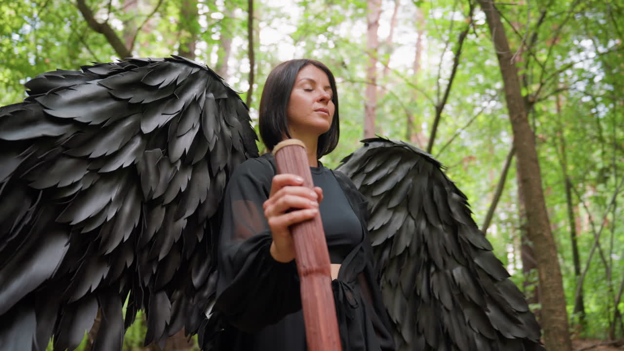Donna determinata vestita di nero con grandi ali scure che cammina in un bosco verde mentre regge un bastone di legno, con la luce del sole che filtra tra gli alberi, che esprime coraggio