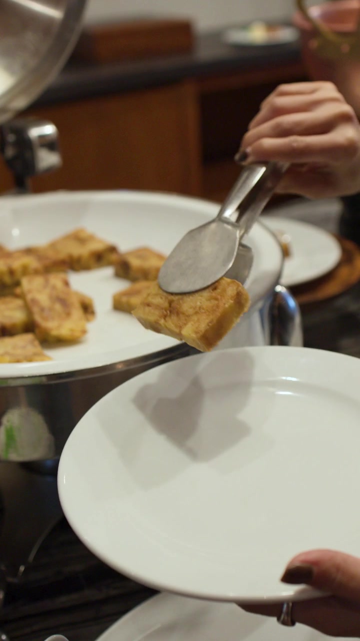 A hand uses metal tongs to pick up Thai custard from a buffet chafing dish