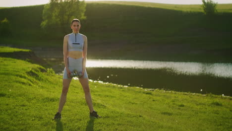 a woman trains with dumbbells in the background of a lake in the park on a sunny day at sunset
