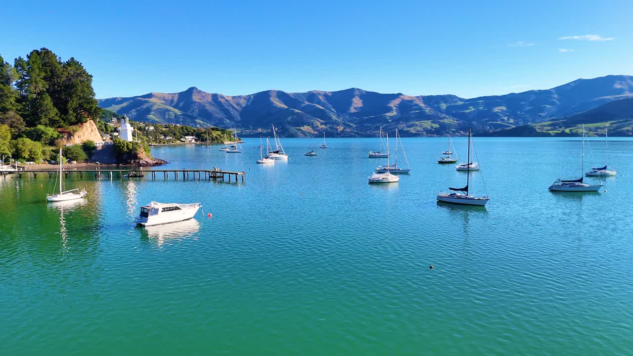 Boats gently float on Akaroa Harbor under clear skies, surrounded by lush hills and tranquil waters, captured in vibrant daylight