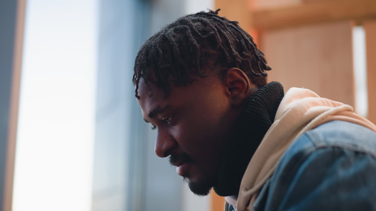 close up view of young man wearing denim jacket and hoodie indoors, moody warm lighting, looking down thoughtfully, textured background, beard stubble and dreadlocks framing face in contemplative pose