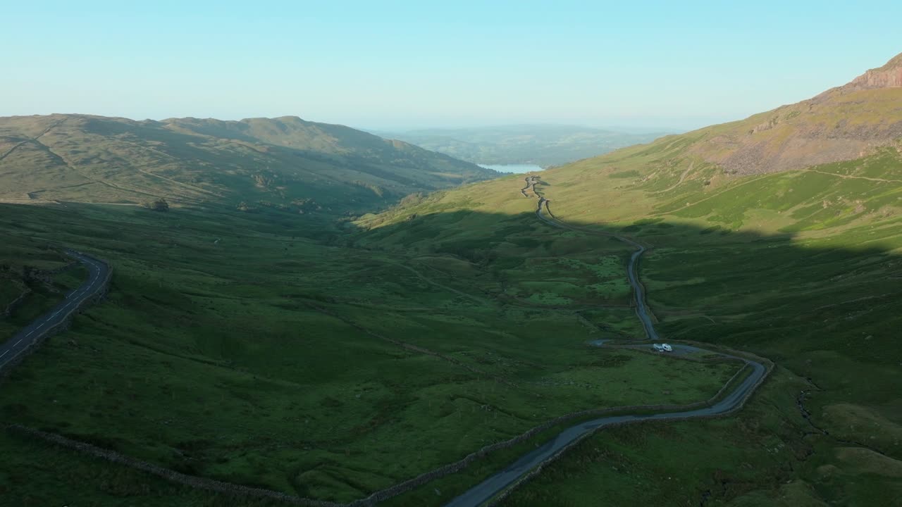 Mountain pass roads in shadowed valley with sunrise light illuminating mountainside. Summer. Kirkstone Pass, Lake District, Cumbria, UK