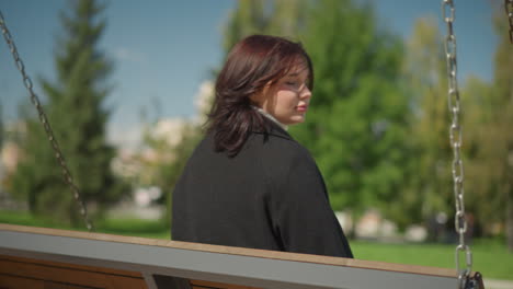 close-up of a woman adjusting her hair while swinging, with sunlight reflecting on her and lush greenery in the background, in public park