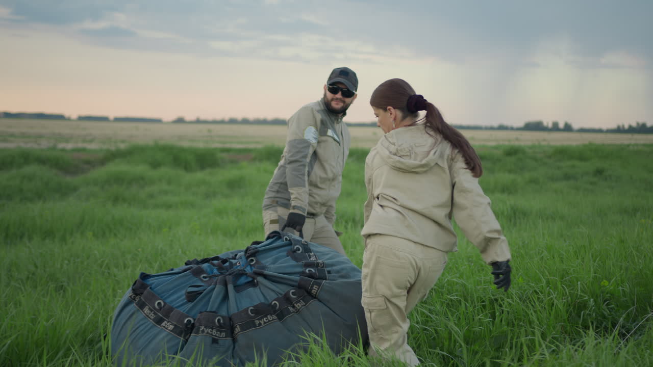 Man and woman dressed in beige uniforms drag heavy blue balloon equipment into wide grassy field under moody sky, pausing with teamwork and focus, surrounded by quiet countryside landscape