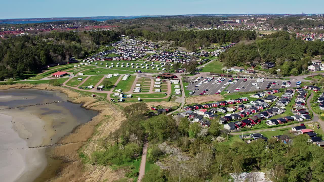 Askimsbadet camping area near Gothenburg, Sweden. Shows rows of cabins, caravans, and parking beside the sandy beach and shoreline, with forested hills and town