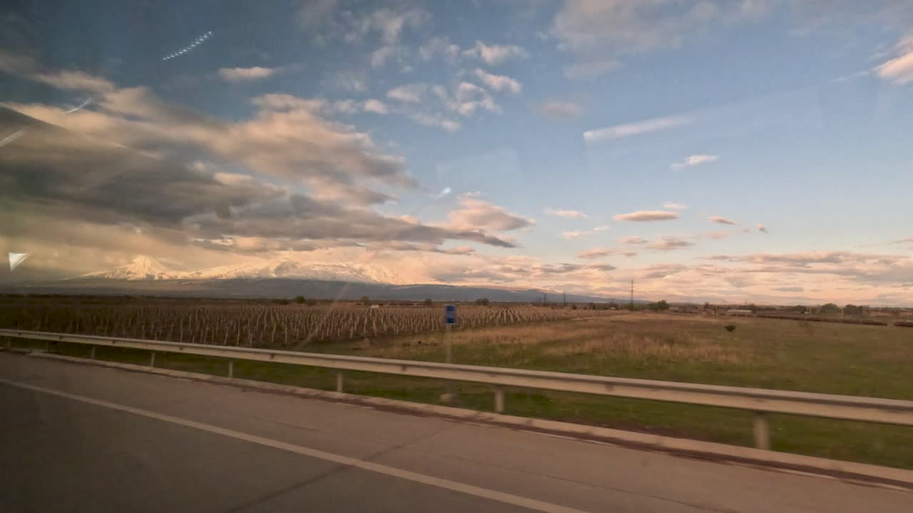 Le montagne si ergono sullo sfondo mentre il sole tramonta dietro le nuvole. I campi si estendono lungo l'autostrada con tralicci e treni visibili