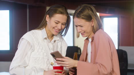 two female colleagues holding drinks and looking something on the mobile phone during a party in the office 1