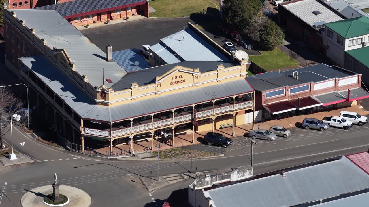 Drone ascends above historic rural town buildings, roundabout, cars, and shops in bright daylight