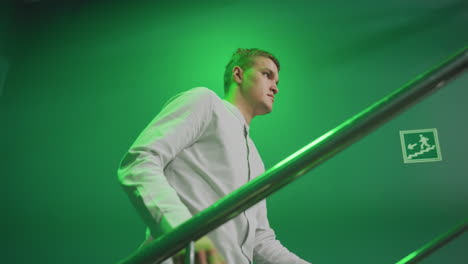 dude with serious face in white shirt and grey trousers walking up stairs with iron rail. green background contrasts with sleek metallic railing, creating a contemplative atmosphere