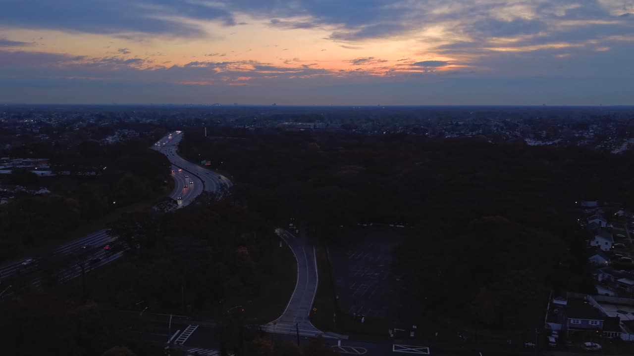 An aerial time lapse over the Southern State Parkway in Valley Stream, NY during a cloudy sunrise. The drone camera, facing the rising sun dolly out and boom down over the residential neighborhood
