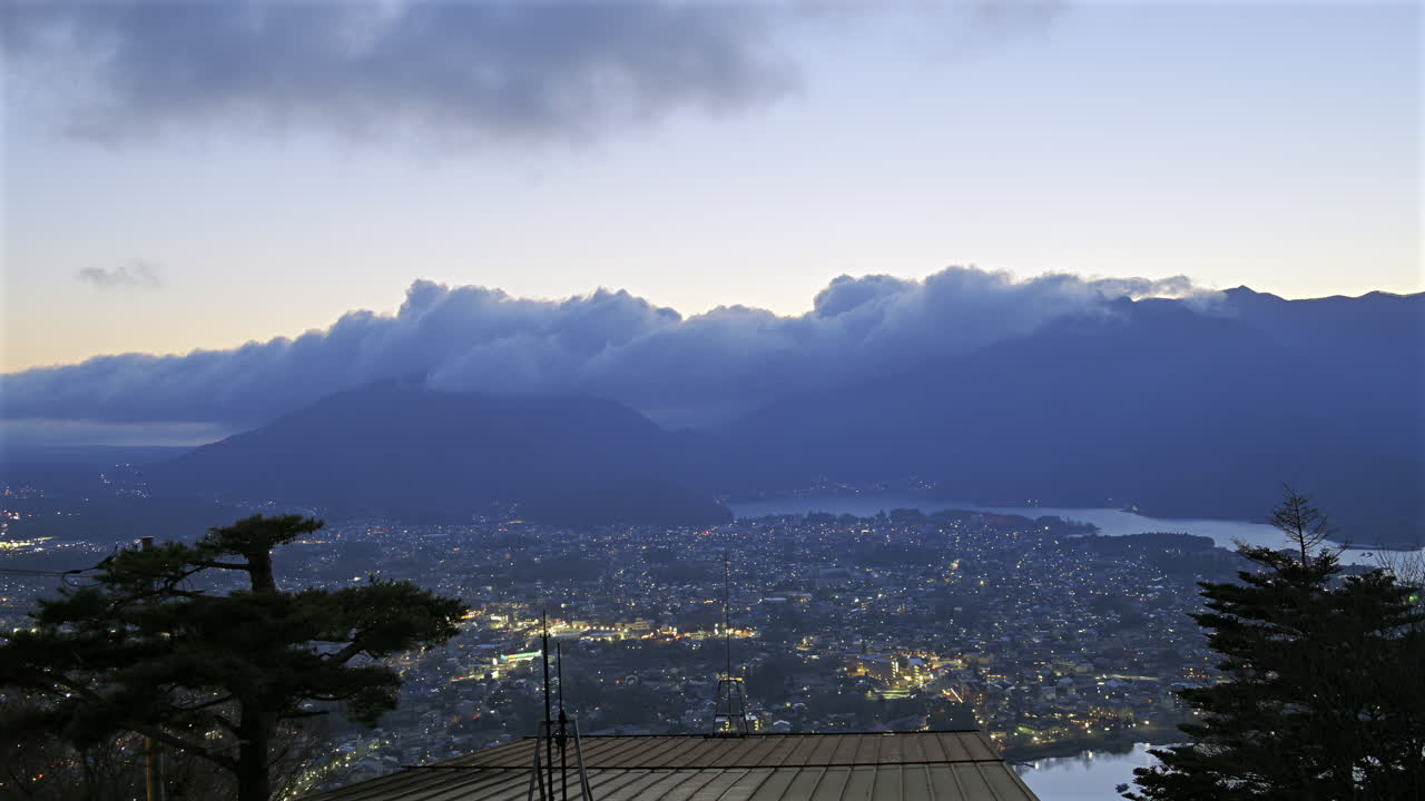 A breathtaking view of Fujikawaguchiko as twilight approaches, with distant mountains draped in clouds. The shimmering lake reflects the changing colors of the sky