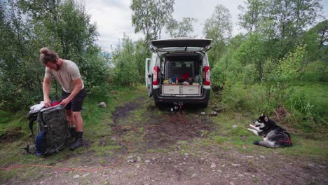 Mann,-Der-Sachen-Mit-Wohnmobil-Packt,-Das-In-Der-Wildnis-Geparkt-Ist,-Und-Alaskischer-Malamute-hund,-Der-Auf-Dem-Boden-Liegt