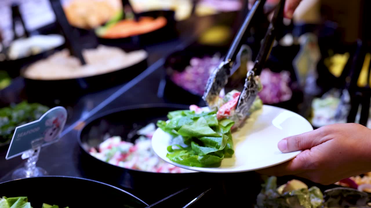 Close-up of diner using tongs to serve fresh salad at colorful buffet bar