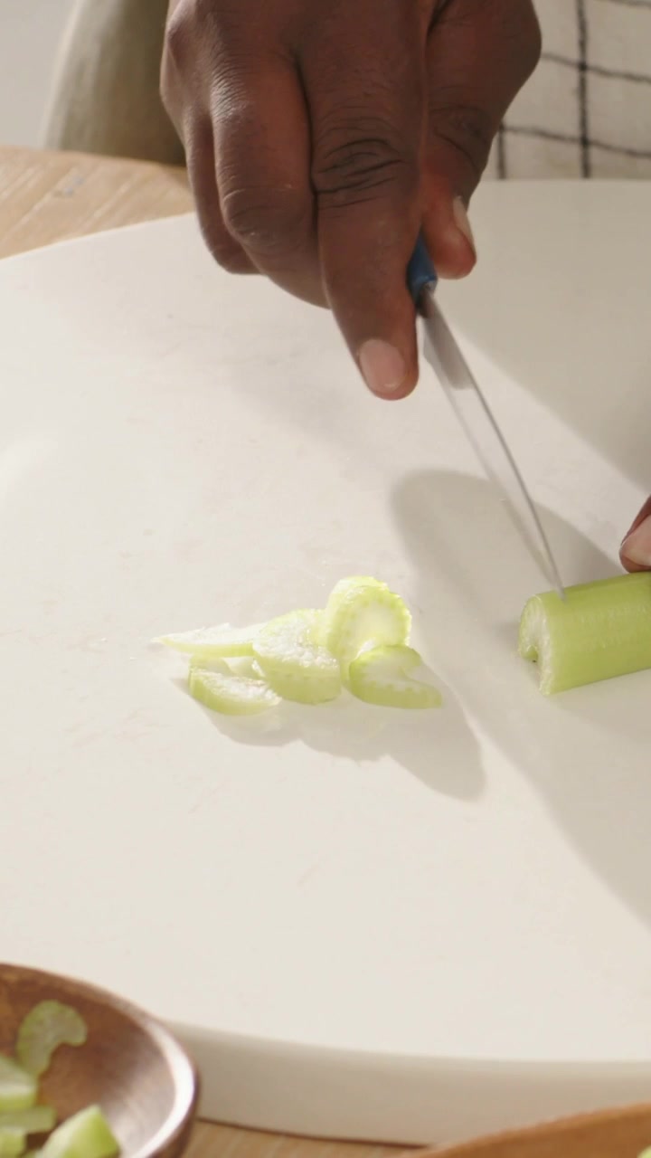 Video verticale: Mani che tengono e tagliano il sedano per l'insalata su un tagliere, con un coltello blu e una ciotola.