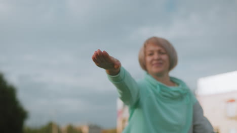 mid aged woman doing exercise with outstretched arm during outdoor fitness routine under cloudy sky wearing teal sportswear smiling confidently in urban park surrounded by greenery