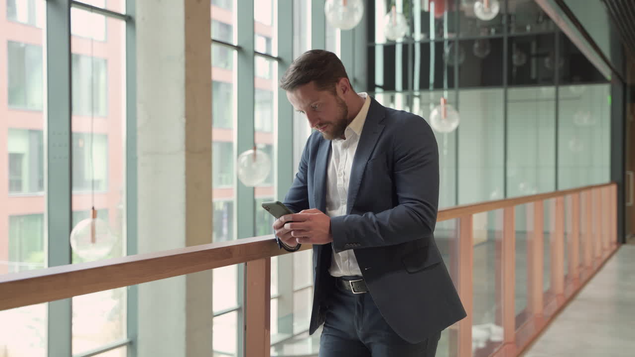 An Attractive Young, Bearded Businessman In A Blue Blazer Consults His Smart Phone Attentively And Writes On It