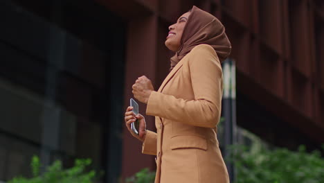 muslim businesswoman wearing hijab standing outside modern office getting good news about job offer or promotion on mobile phone 3