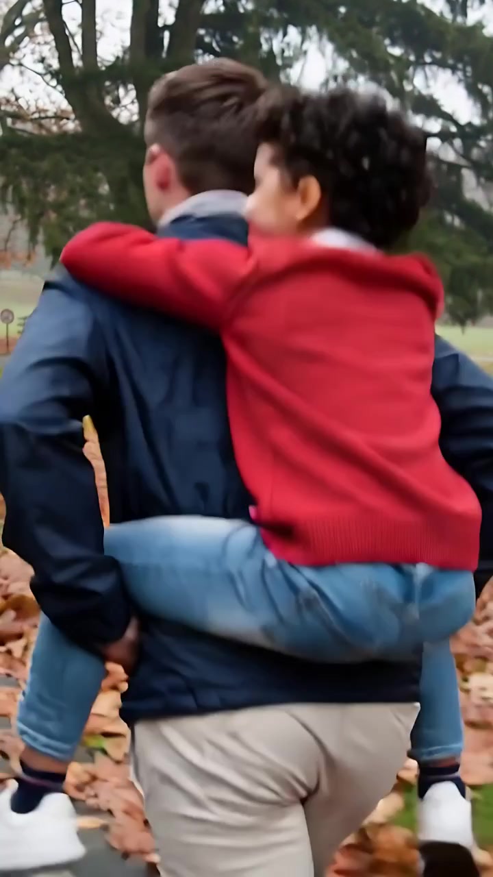 Video verticale: Uomo adulto che porta un bambino in spalla lungo un sentiero con foglie, felpa con cappuccio rossa.