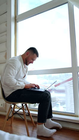 positive caucasian man sitting on the chair works on laptop. smiling man typing on his gadget near the window. vertical video.