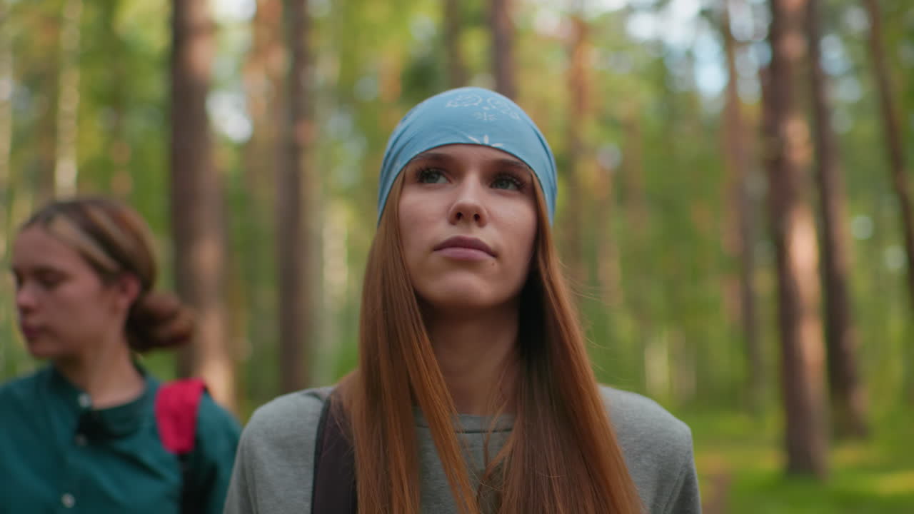 mujer con bandana azul caminando por el sendero del bosque con una sonrisa sutil, emanando confianza tranquila, mientras su amiga con camisa verde mira a su alrededor pacíficamente en un fondo borroso suave en medio de árboles altos