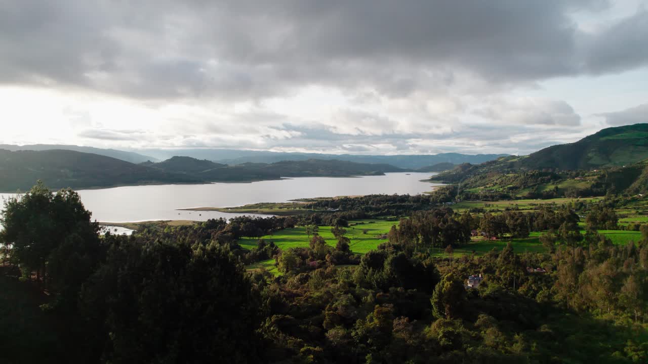 Drone zoom out from Guatavita, Colombia, showing Tominé Reservoir and the surrounding green landscape under dramatic cloudy sky