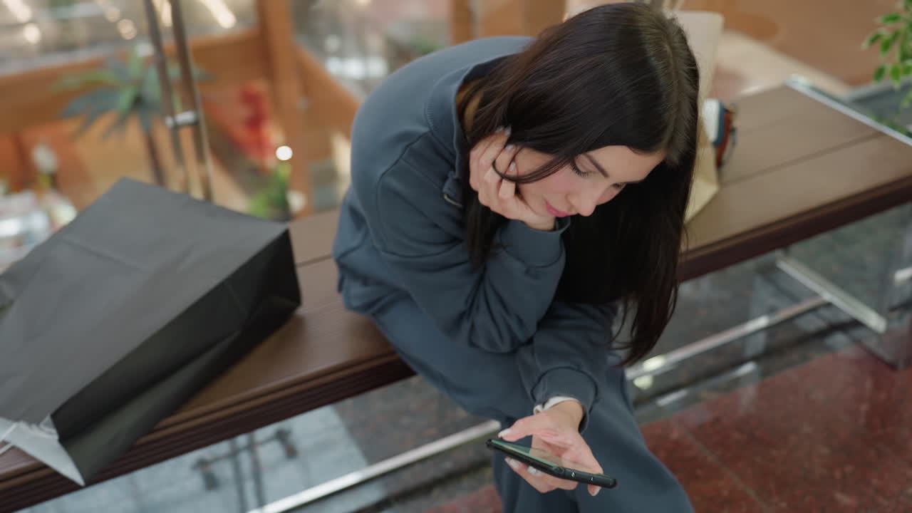Young woman with long dark hair in casual dark tracksuit sitting on bench in indoor public space, leaning forward with hand on chin, focused on smartphone screen