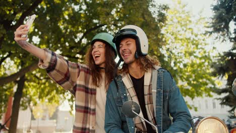 happy couple, a brunette girl in a plaid shirt takes a selfie using a phone together with her boyfriend with long curly hair in a moped helmet and a denim shirt who sits on a moped in a sunny summer park