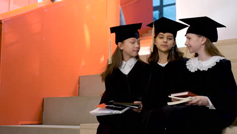 three happy little girls in cap and gown talking and holding their notebooks while sitting on stairs at the preschool graduation ceremony