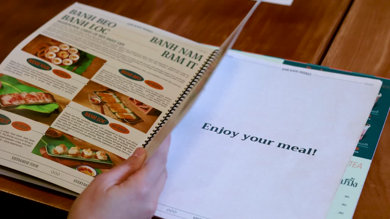 En person bladrer i en spiralindbundet vietnamesisk madmenu på et træbord i Thailand.