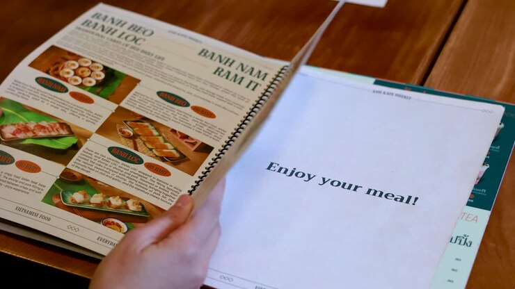 Una persona sfoglia un menu di cibo vietnamita rilegato a spirale su un tavolo di legno in Thailandia.