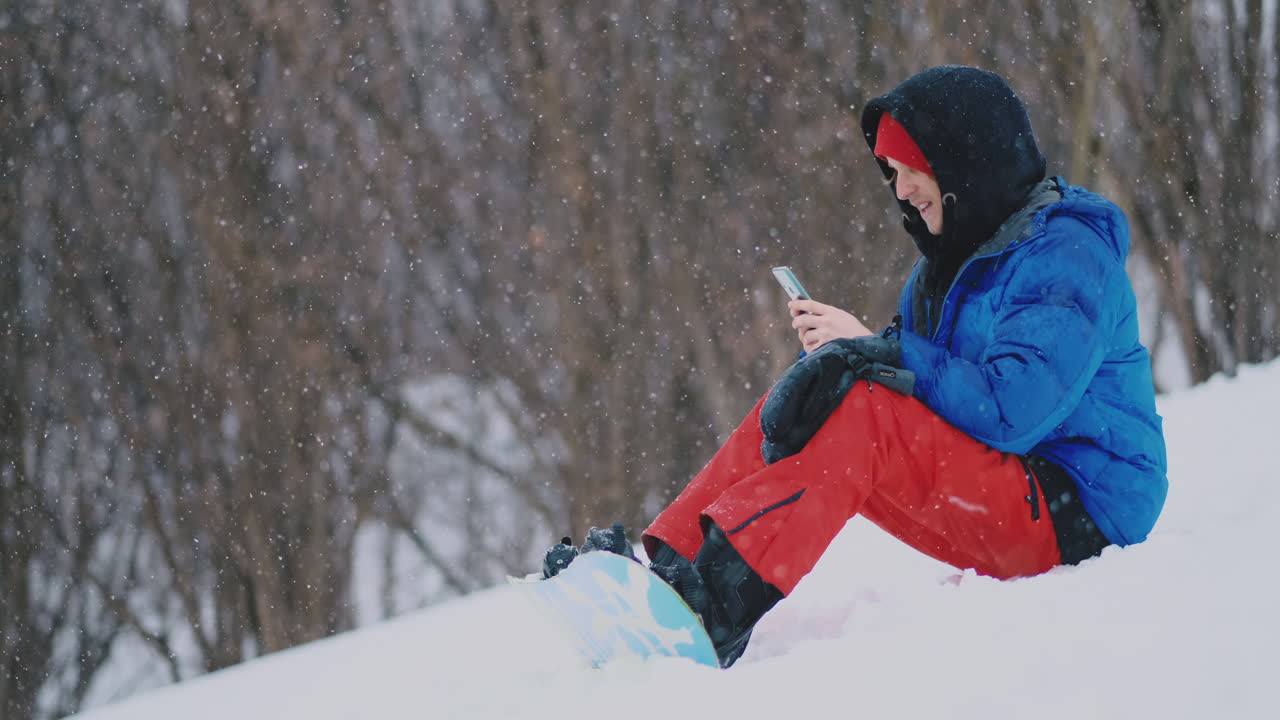 A male snowboarder sitting on the snow takes photos on the phone of a beautiful resort landscape for social networks. Resort blogger