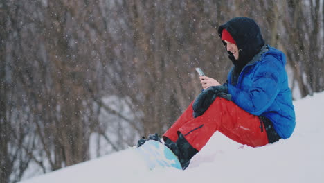 a male snowboarder sitting on the snow takes photos on the phone of a beautiful resort landscape for social networks. resort blogger