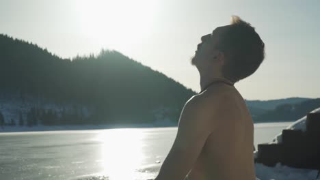 hombre sin camisa sentado afuera meditando cerca del lago de montaña congelado en invierno con nieve en el suelo