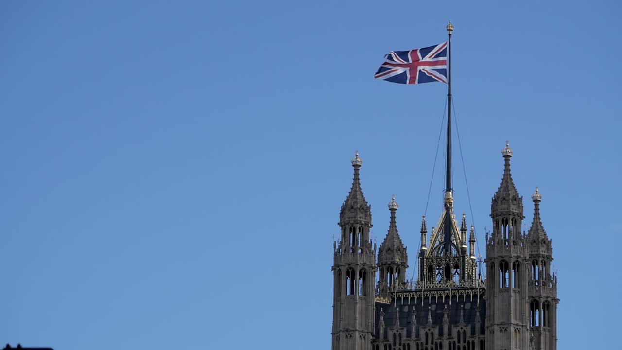 Top of Victoria Tower, Palace of Westminster in London, United Kingdom. Union Jack, flag flies on sky. Square tower. Perpendicular Gothic stonework golden ornaments. Holds Parliamentary Archives.
