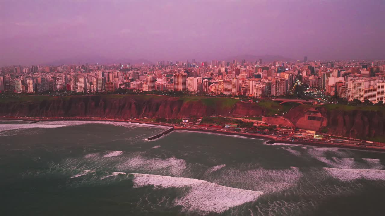 Afterglow sunset Miraflores coastal seaside aerial drone harbor view Lima Peru Makaha Barranco beach pink purple clear skies hazy cityscape skyscraper buildings ocean waves cars traffic forward pan up