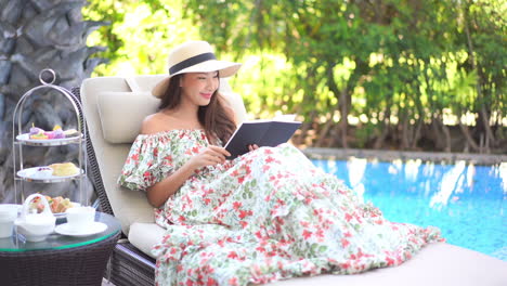 elegant young asian woman in summer dress sitting by the pool reading the book