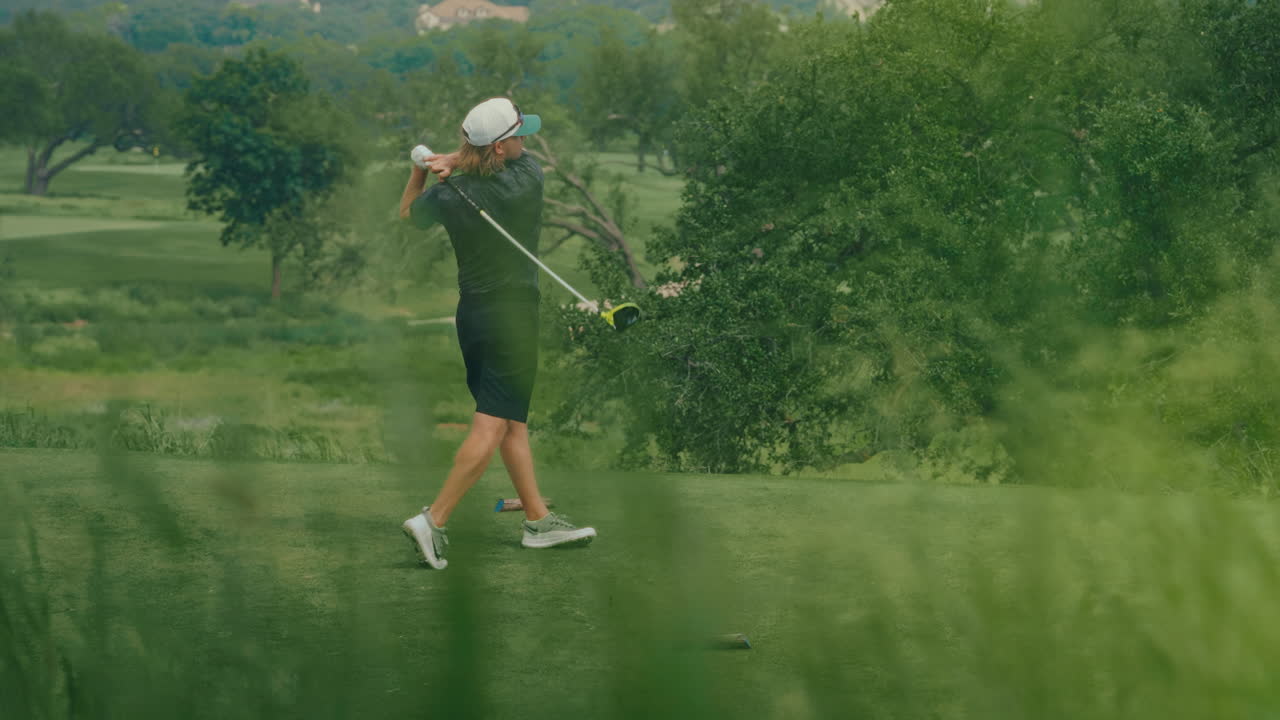 Male golfer hits a powerful drive in slow motion, framed by lush green landscape. The clip captures the smooth swing and natural beauty of the golf course in a vibrant outdoor setting.