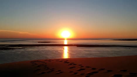 Drohnenvideo-Eines-Wunderschönen-Orangefarbenen-Sonnenuntergangs-Auf-Einer-Sanddüne-Mit-Meereshintergrund,-Sand-Hat-Fußabdrücke-Und-Sieht-Aus-Wie-Der-Reine-Himmel