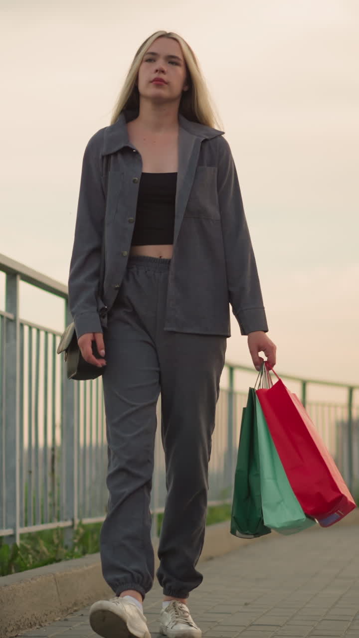 dama de ropa gris caminando por el ferrocarril, sosteniendo bolsas de compras de colores y bolso de mano negro, parece agotada, con una vista borrosa del lejano puente sobre el río en el fondo